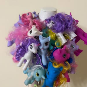 May include: A collection of small plastic pony figurines in various colors including pink, purple, blue, and white, featuring rooted synthetic hair in shades of yellow, pink, and purple. The ponies are arranged in a dense cluster on a white base.