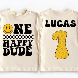 Könnte beinhalten: Cremefarbenes Kleinkind-T-Shirt mit dem Text "ONE HAPPY DUDE" und einem Smiley-Gesicht. Auf der Rückseite steht "LUCAS" und die Zahl 1 mit Smiley-Gesichtern. Ein lustiges Geburtstags-T-Shirt im Retro-Stil.