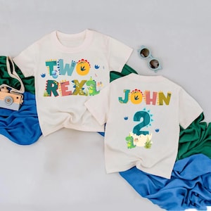 May include: Two white t-shirts with dinosaur-themed graphics. One shirt says "TWO REX" in colorful letters. The other shirt says "JOHN" and "2" with a dinosaur hatching from an egg. Sunglasses are on the side.