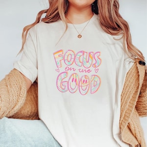May include: A cream-coloured t-shirt featuring the phrase "Focus on the Good" in a vibrant, swirly font. The design incorporates hearts and a smiley face. This is a casual, everyday wear item.