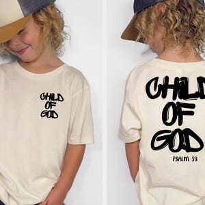 May include: Cream-colored t-shirt with the words "CHILD OF GOD" in black, stylized lettering on the front and back. The back also includes "PSALM 23". The shirt is worn by a child wearing a baseball cap.