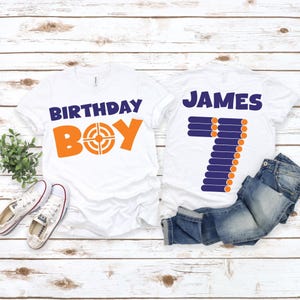 May include: Two white t-shirts with orange and blue text. One shirt says "BIRTHDAY BOY" with a target graphic. The other says "JAMES 7" with a dart design. The shirts are on a white wood surface.