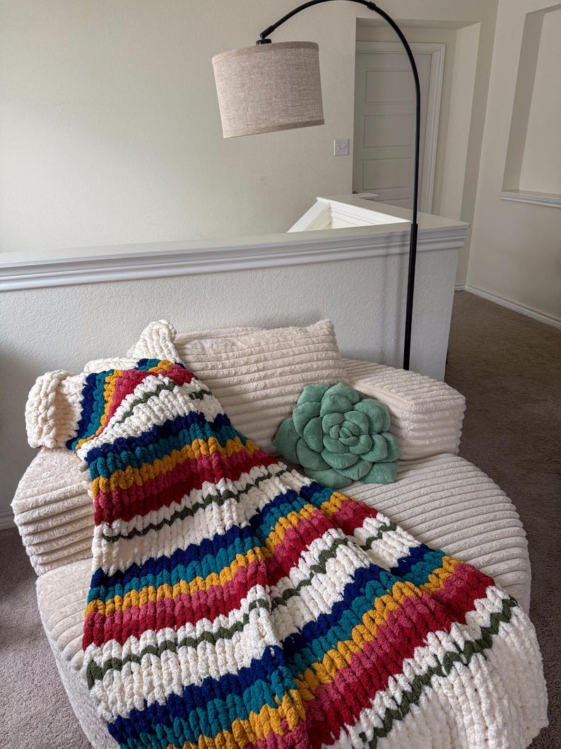 Puede incluir: Una colorida manta de punto grueso en tonos arco&iacute;ris, incluyendo rojo, azul, amarillo y verde, sobre una silla color crema. Tambi&eacute;n se ven una almohada verde con forma de suculenta y una l&aacute;mpara beige con una pantalla neutra.