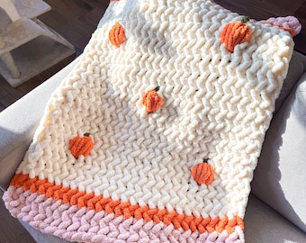 Puede incluir: Una manta de punto grueso en color crema con pequeñas aplicaciones de calabazas naranjas en relieve repartidas por la superficie. La manta tiene un borde ancho en la parte inferior con rayas horizontales de color naranja y rosa claro.