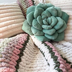 Puede incluir: Un coj&iacute;n en forma de suculenta verde salvia descansa sobre una almohada crema acanalada. Una manta de punto grueso en tonos rosa, verde y crema est&aacute; drapeada debajo. Tambi&eacute;n se ven otras almohadas en el fondo.