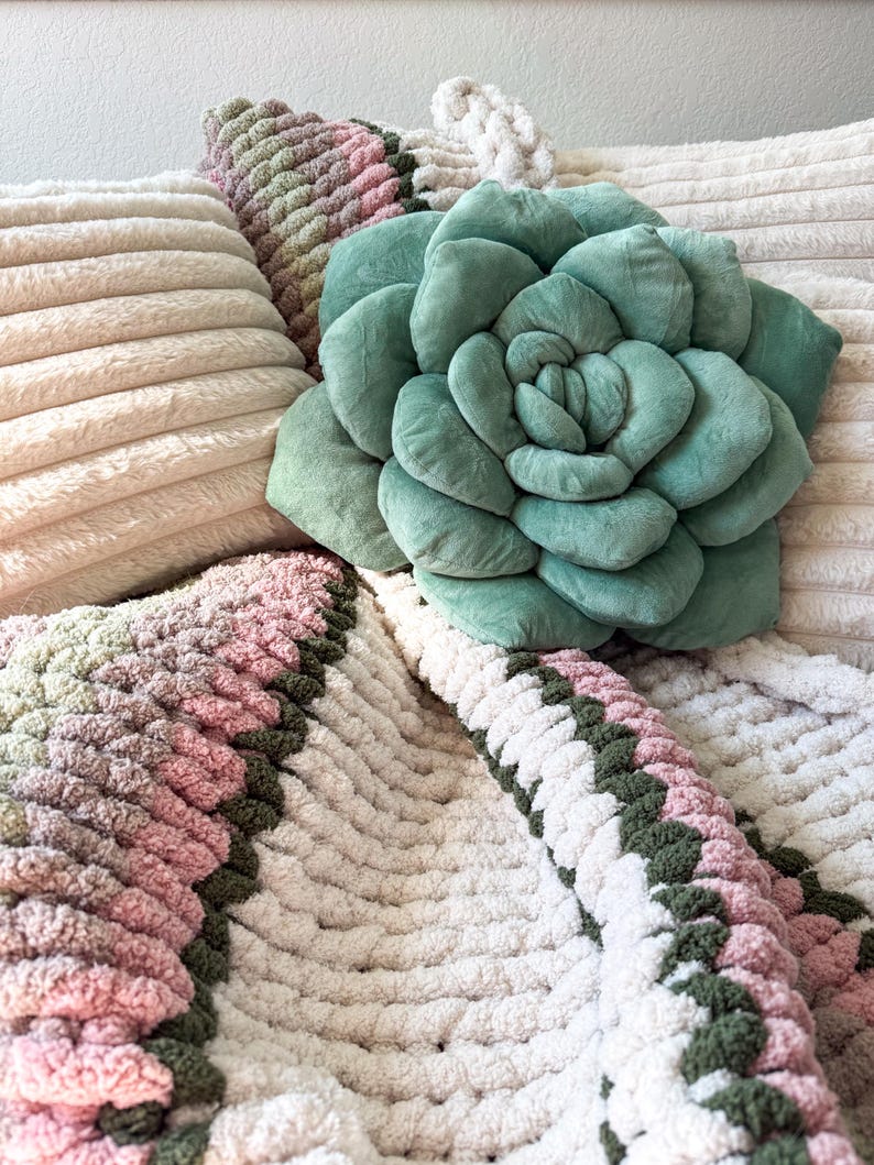 Puede incluir: Un coj&iacute;n en forma de suculenta verde salvia descansa sobre una almohada crema acanalada. Una manta de punto grueso en tonos rosa, verde y crema est&aacute; drapeada debajo. Tambi&eacute;n se ven otras almohadas en el fondo.
