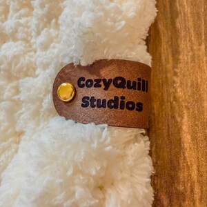 Puede incluir: Primer plano de un textil cremoso y esponjoso con una etiqueta de cuero marr&oacute;n. La etiqueta est&aacute; asegurada con un bot&oacute;n dorado y presenta el texto "CozyQuill Studios" en letras marr&oacute;n oscuro. El fondo es una superficie de madera.