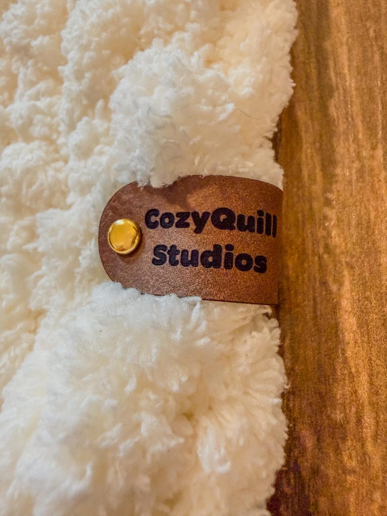 Puede incluir: Primer plano de un textil cremoso y esponjoso con una etiqueta de cuero marr&oacute;n. La etiqueta est&aacute; asegurada con un bot&oacute;n dorado y presenta el texto "CozyQuill Studios" en letras marr&oacute;n oscuro. El fondo es una superficie de madera.