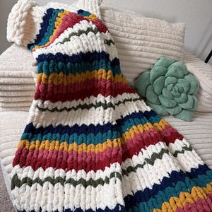 Puede incluir: Una manta de punto grueso con un vibrante dise&ntilde;o de arco&iacute;ris. La manta presenta rayas alternas blancas, rojas, amarillas, verdes, turquesas y azul marino. La suave y texturizada manta est&aacute; sobre una superficie color crema, con una almohada verde en forma de suculenta en el fondo.
