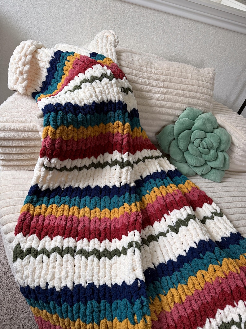 Puede incluir: Una manta de punto grueso con un vibrante dise&ntilde;o de arco&iacute;ris. La manta presenta rayas alternas blancas, rojas, amarillas, verdes, turquesas y azul marino. La suave y texturizada manta est&aacute; sobre una superficie color crema, con una almohada verde en forma de suculenta en el fondo.