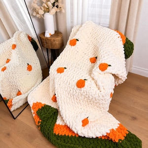 Könnte beinhalten: Eine grob gestrickte Decke aus cremefarbenem Garn mit kleinen orangefarbenen Kürbismotiven und grünen Stielen. Die Decke hat am unteren Rand eine breite Bordüre aus abwechselnd orangefarbenem und dunkelgrünem Garn mit strukturierter Optik.