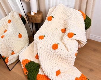 Puede incluir: Una manta de punto grueso en color crema con pequeños motivos de calabazas naranjas con tallos verdes. La manta tiene un borde ancho de hilo naranja y verde oscuro en la parte inferior, creando una apariencia texturizada y suave al tacto.