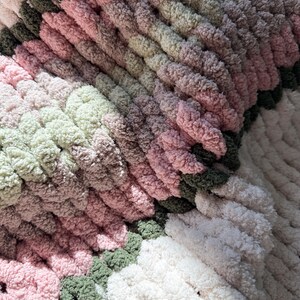 Puede incluir: Primer plano de una manta de punto grueso hecha a mano. La manta presenta un dise&ntilde;o de bloques de color con tonos de rosa, verde y blanco. El hilo grueso crea una apariencia texturizada y acogedora, ideal para la decoraci&oacute;n del hogar.