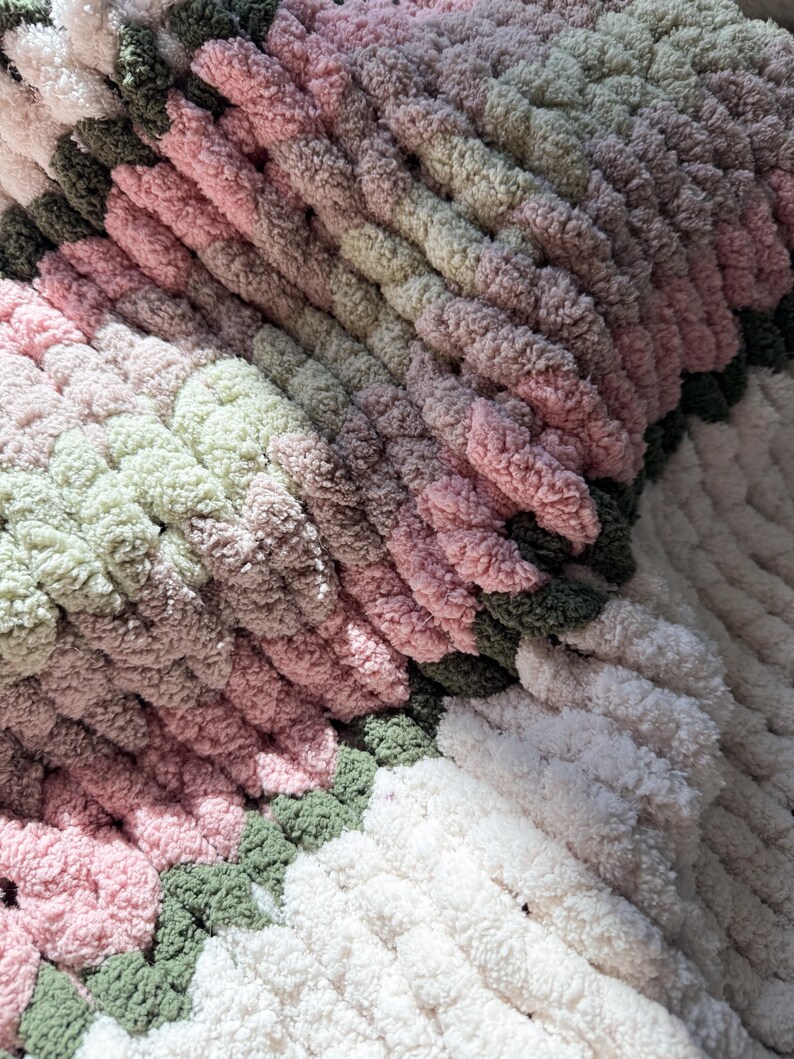 Puede incluir: Primer plano de una manta de punto grueso hecha a mano. La manta presenta un dise&ntilde;o de bloques de color con tonos de rosa, verde y blanco. El hilo grueso crea una apariencia texturizada y acogedora, ideal para la decoraci&oacute;n del hogar.