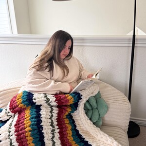 Puede incluir: Una manta de punto grueso color crema, con rayas rojas, turquesas, amarillas y azules. La manta est&aacute; sobre una persona leyendo un libro. La manta est&aacute; hecha de hilo grueso y suave, ideal para relajarse en el sof&aacute;.