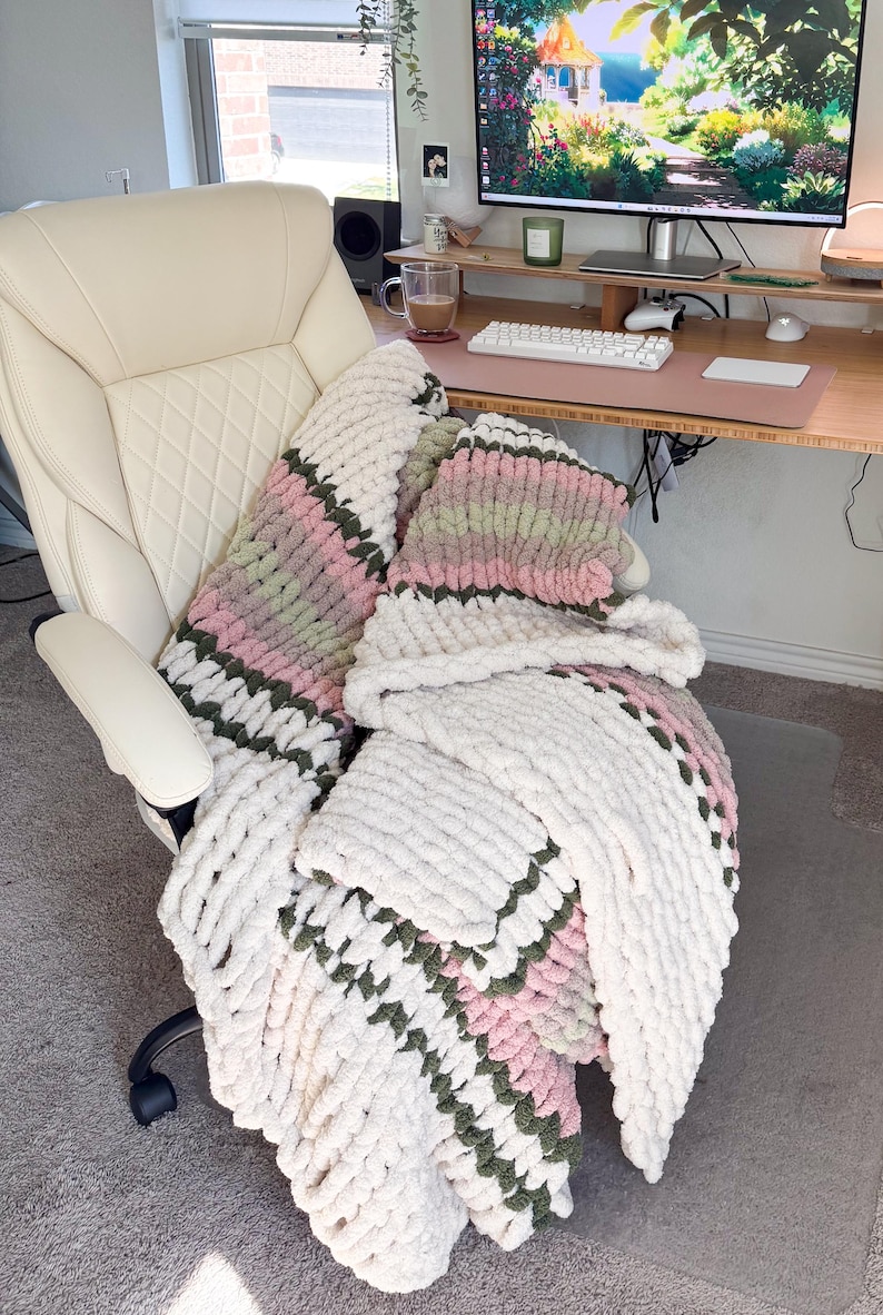 Puede incluir: Una manta de punto grueso en blanco, rosa y verde sobre una silla de oficina color crema. El hilo grueso de la manta crea una apariencia texturizada y acogedora. La silla est&aacute; frente a un escritorio con una computadora.