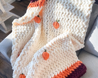 Puede incluir: Una manta de punto grueso hecha de hilo color crema, con pequeñas aplicaciones de calabazas naranjas con tallos verdes. La manta presenta anchas franjas horizontales de color naranja y burdeos oscuro en los bordes.
