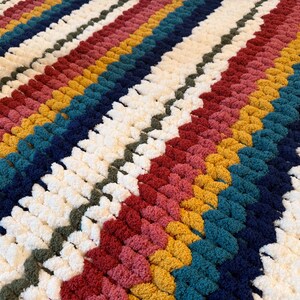 Puede incluir: Una manta hecha a mano con un dise&ntilde;o de rayas. La manta presenta filas de varios colores, incluyendo blanco, rojo, amarillo, turquesa y azul marino. La textura parece suave y afelpada, lo que sugiere un art&iacute;culo c&aacute;lido y acogedor.