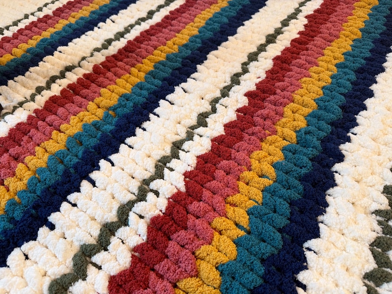 Puede incluir: Una manta hecha a mano con un dise&ntilde;o de rayas. La manta presenta filas de varios colores, incluyendo blanco, rojo, amarillo, turquesa y azul marino. La textura parece suave y afelpada, lo que sugiere un art&iacute;culo c&aacute;lido y acogedor.