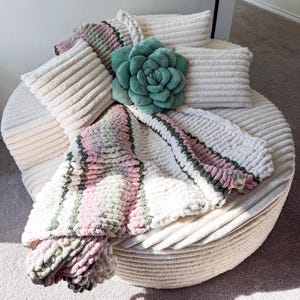 Puede incluir: Un otom&aacute;n redondo y cremoso est&aacute; adornado con cojines y una manta. Los cojines son rectangulares y con textura de pana. Una gran almohada en forma de flor, de color verde azulado, a&ntilde;ade un toque de color. La manta presenta rayas rosas, verdes y blancas.