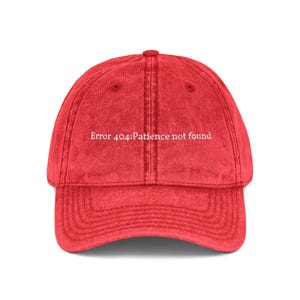 May include: A red baseball cap with a curved brim. The cap features the text "Error 404: Patience not found" in white, centered on the front. The hat has a vintage, worn look.
