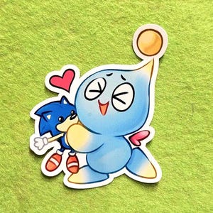 May include: A sticker featuring a light blue Chao character hugging a blue Sonic the Hedgehog. The Chao has a yellow orb on its head and a pink bow. A red heart is above them. The sticker is on a green background.
