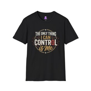 May include: A black short-sleeved t-shirt featuring a circular graphic design with the text "THE ONLY THING I CAN CONTROL is Me" in white and gold lettering. The design includes small stars and radiating lines, centred on the chest of the shirt.