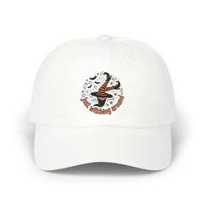 Op de afbeelding: Een witte baseballpet met een afbeelding van heksenbenen in oranje en zwart gestreepte kousen die uit een zwarte puntmuts steken. Het ontwerp bevat kleine vleermuizen, sterren en de tekst Just witching around in oranje letters.