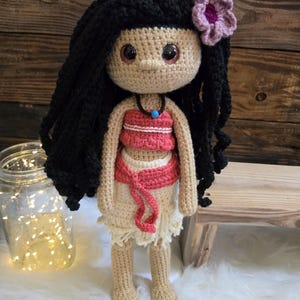 May include: A handcrafted crochet doll with long black yarn hair, a pink flower accessory, and large brown eyes. The doll wears a coral-colored top, a matching coral waist tie, and a cream-colored skirt, accented by a dark necklace with a blue bead.
