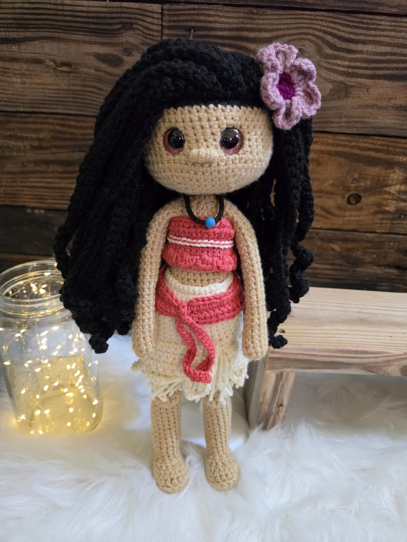 May include: A handcrafted crochet doll with long black yarn hair, a pink flower accessory, and large brown eyes. The doll wears a coral-colored top, a matching coral waist tie, and a cream-colored skirt, accented by a dark necklace with a blue bead.
