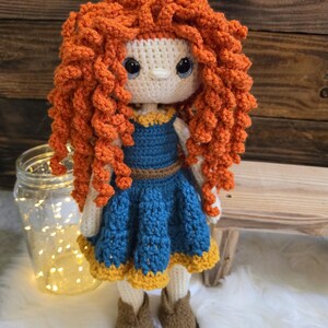 May include: A handmade crochet doll featuring voluminous, curly orange yarn hair, large dark eyes, and a cream-colored face. The doll wears a blue dress with a brown belt and a yellow hem, paired with brown crochet boots, crafted with intricate yarn textures.