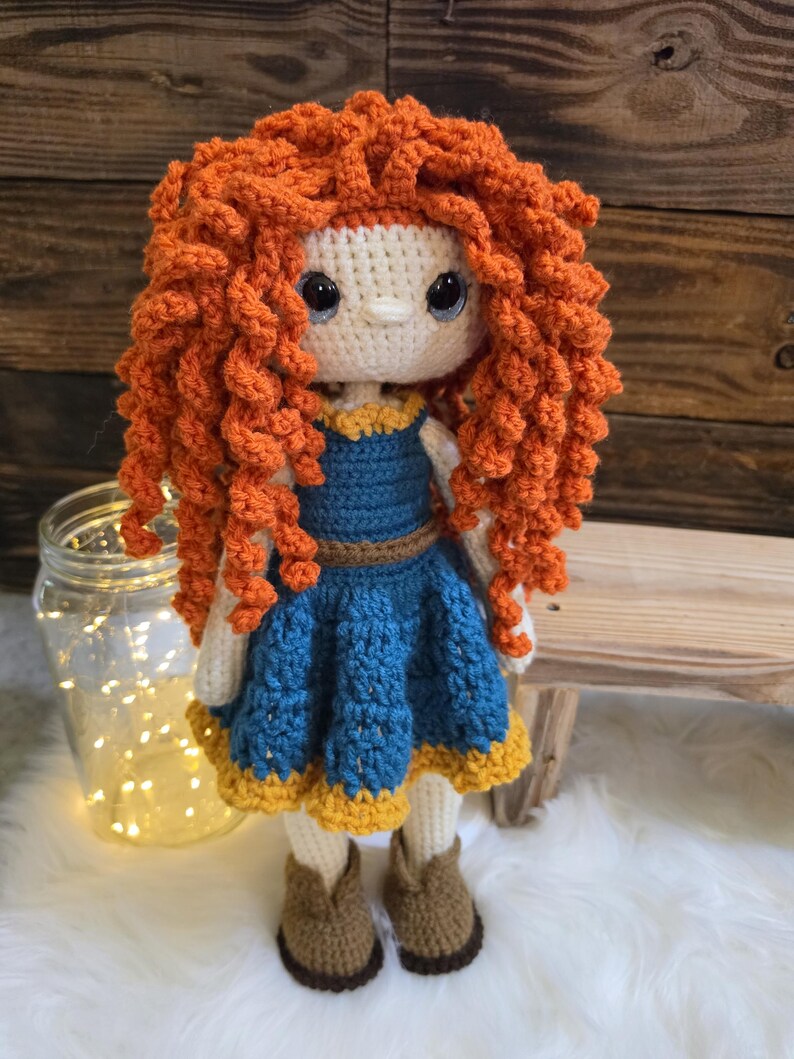 May include: A handmade crochet doll featuring voluminous, curly orange yarn hair, large dark eyes, and a cream-colored face. The doll wears a blue dress with a brown belt and a yellow hem, paired with brown crochet boots, crafted with intricate yarn textures.