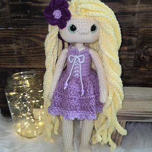 May include: A handmade crochet doll featuring long, wavy yellow yarn hair adorned with a purple flower. The doll has large green eyes and wears a lavender dress with a white lace-up bodice detail, featuring two small purple beads at the ends of the strings.