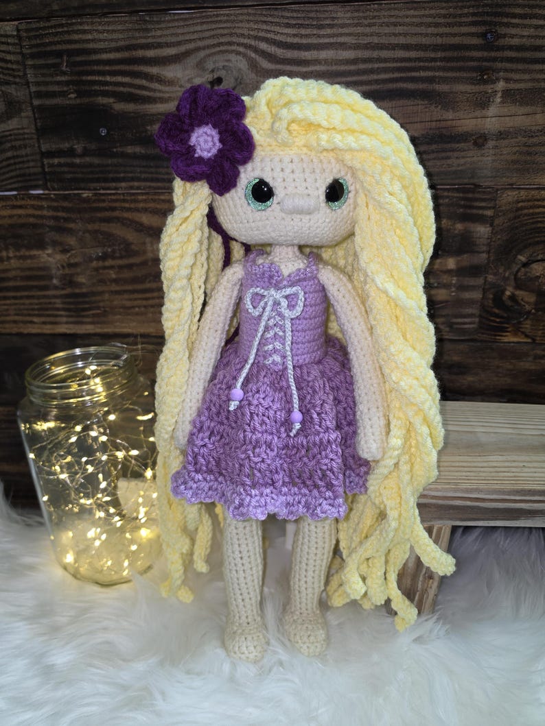 May include: A handmade crochet doll featuring long, wavy yellow yarn hair adorned with a purple flower. The doll has large green eyes and wears a lavender dress with a white lace-up bodice detail, featuring two small purple beads at the ends of the strings.