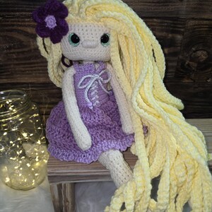 May include: A handmade crochet doll with long, wavy yellow yarn hair, a purple flower accessory, and a light beige complexion. The doll wears a purple crocheted dress featuring a white lace-up bodice detail and has large, round, dark-rimmed green eyes.