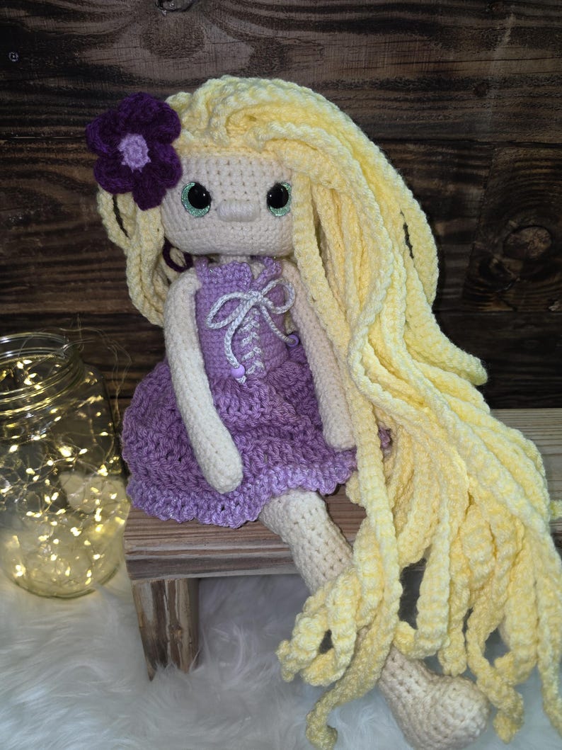 May include: A handmade crochet doll with long, wavy yellow yarn hair, a purple flower accessory, and a light beige complexion. The doll wears a purple crocheted dress featuring a white lace-up bodice detail and has large, round, dark-rimmed green eyes.