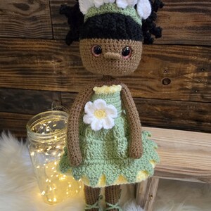 May include: A handcrafted crochet doll with brown skin, black curly hair, and a white floral headband. The doll wears a light green dress featuring a white flower appliqu&eacute; on the chest, a yellow underskirt, and matching green lace-up ballet slippers.