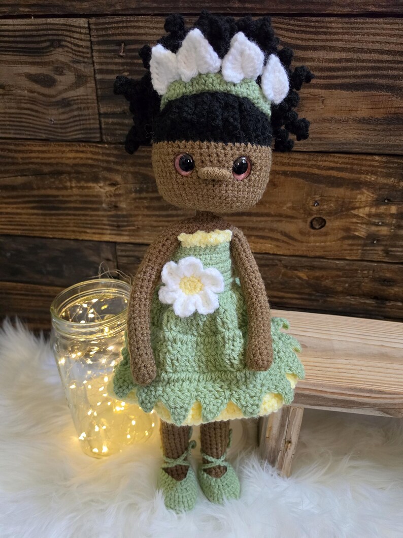 May include: A handcrafted crochet doll with brown skin, black curly hair, and a white floral headband. The doll wears a light green dress featuring a white flower appliqu&eacute; on the chest, a yellow underskirt, and matching green lace-up ballet slippers.