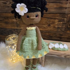 May include: A handmade crochet doll with dark brown skin, curly black hair, and a white flower accessory. The doll wears a light green dress with a yellow underskirt and matching green lace-up shoes. A separate green headband with white petals is included.