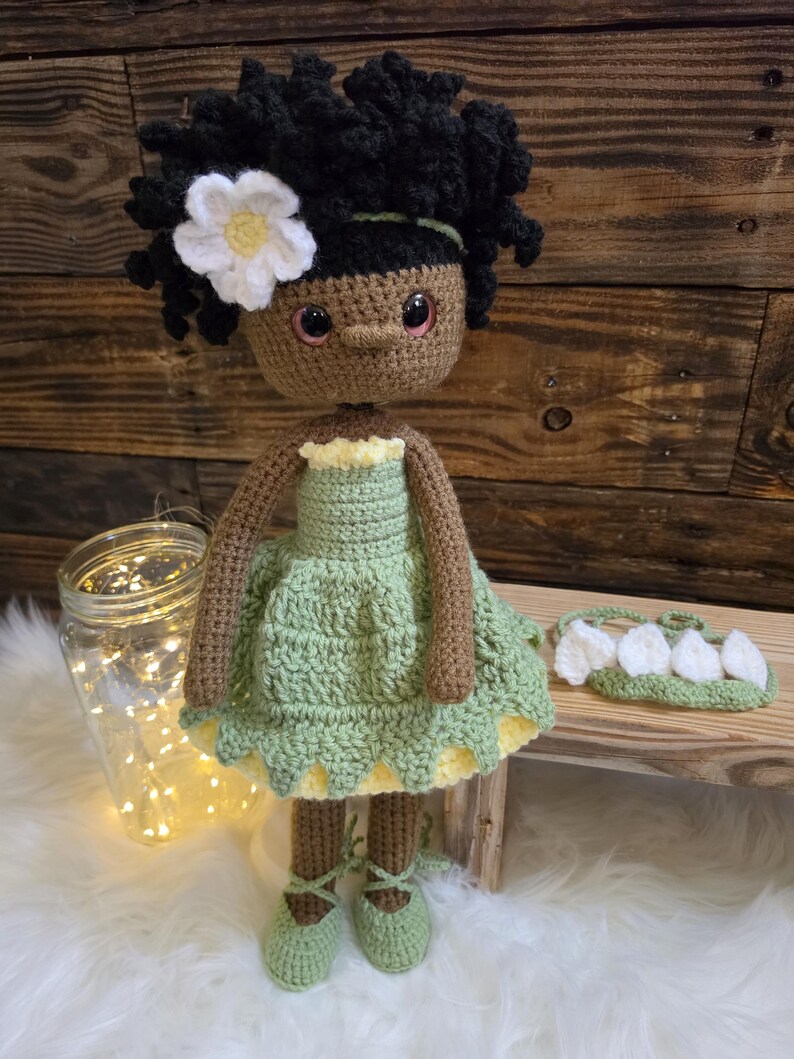 May include: A handmade crochet doll with dark brown skin, curly black hair, and a white flower accessory. The doll wears a light green dress with a yellow underskirt and matching green lace-up shoes. A separate green headband with white petals is included.
