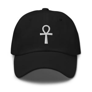 May include: A black baseball cap featuring a white embroidered ankh symbol centered on the front panel. The cap has a structured crown, a curved brim, and a button at the top. The embroidery shows a clear, detailed texture of the Egyptian cross.