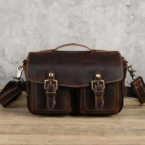 Puede incluir: Bolso mensajero de cuero marrón oscuro con dos bolsillos delanteros, hebillas de latón y un asa superior. El bolso tiene una correa para el hombro. El cuero tiene un aspecto envejecido. Diseñado para transportar objetos.