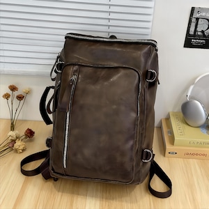 Könnte beinhalten: Ein brauner Lederrucksack mit mehreren Fächern und silbernen Reißverschlüssen. Die Tasche hat verstellbare Träger und einen oberen Griff. Das Leder hat einen Used-Look. Der Rucksack steht auf einer Holzoberfläche.