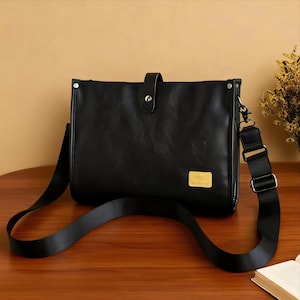 Könnte beinhalten: Schwarze Umhängetasche mit einem langen, verstellbaren Riemen. Die Tasche hat einen Klappenverschluss mit einem Knopf und ein kleines goldfarbenes rechteckiges Emblem auf der Vorderseite. Die Tasche ist aus glattem, lederähnlichem Material.