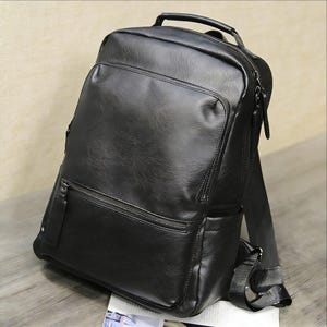 Può includere: Uno zaino in pelle nera con più scomparti con cerniera e una maniglia superiore. La borsa ha spallacci regolabili e una superficie strutturata. Lo zaino è appoggiato su una superficie di legno.