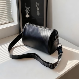 Könnte beinhalten: Schwarze Umhängetasche in zylindrischer Form mit einem langen, verstellbaren Riemen. Sie hat einen silbernen Reißverschluss und Metallbeschläge. Die Tasche ist aus glattem, lederähnlichem Material gefertigt.