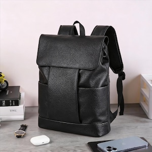 Peut inclure: Sac à dos noir avec fermeture à rabat et deux poches avant. Le sac à dos a des bretelles réglables et une poignée supérieure. Le matériau a un aspect texturé. Le sac est posé sur une table.