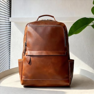 Könnte beinhalten: Ein brauner Lederrucksack mit einem oberen Griff und mehreren Fächern. Der Rucksack hat eine rechteckige Form mit einer Frontklappe und Seitentaschen. Die Reißverschlüsse und die Hardware sind silberfarben. Die Tasche steht auf einer weißen Oberfläche.