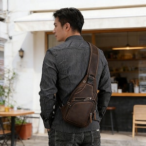 Könnte beinhalten: Eine braune Leder-Umhängetasche mit einem einzelnen Schulterriemen. Die Tasche hat mehrere Reißverschlussfächer und einen Schnallenverschluss. Die Person trägt ein dunkelgraues gestreiftes Hemd und dunkle Jeans.