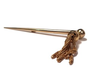 May include: A gold-toned metal pin with a tassel. The pin is long and slender with a rounded end. The tassel is made of braided gold-toned metal.